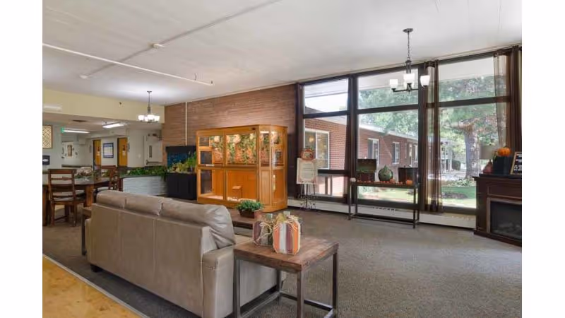 A spacious living room area in a senior living facility with a beige couch facing a large wooden display cabinet and an aquarium. There is a wooden side table with decorative items next to the couch. Large windows on the right side let in natural light and offer a view of the outdoor garden area. The room has carpeted flooring and a chandelier hanging from the ceiling. In the background, there is a dining area with wooden chairs and tables.