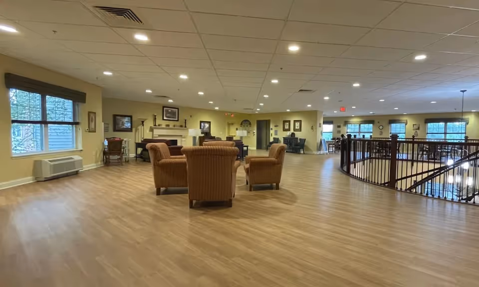 Spacious senior living common area with wood floors, grouped armchairs around a fireplace, windows, and a railing overlooking a lower level.