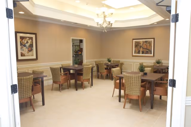 A dining room with several square wooden tables, each surrounded by four cushioned chairs with patterned upholstery. The room has beige walls with white wainscoting and two framed paintings hanging on the walls. A chandelier hangs from a recessed ceiling with soft lighting. There are small potted plants on some tables, and an open doorway leads to another room.