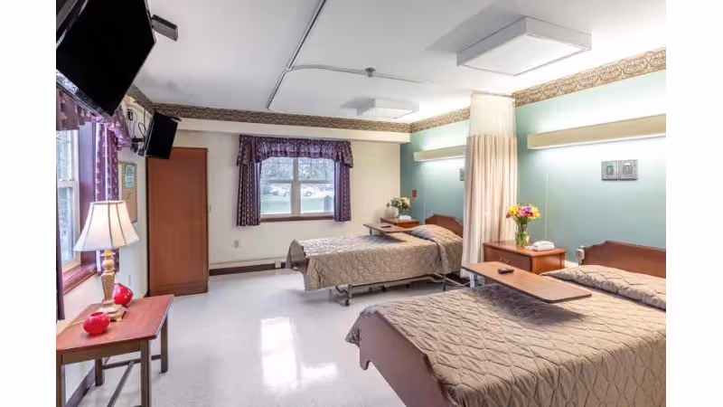 A well-lit room with two single beds covered in beige quilted bedspreads. Each bed has a wooden headboard and a bedside table with a lamp and flowers. There is a window with purple curtains, a wooden wardrobe, and a TV mounted on the wall. The room has a light-colored floor and walls with a decorative border near the ceiling.