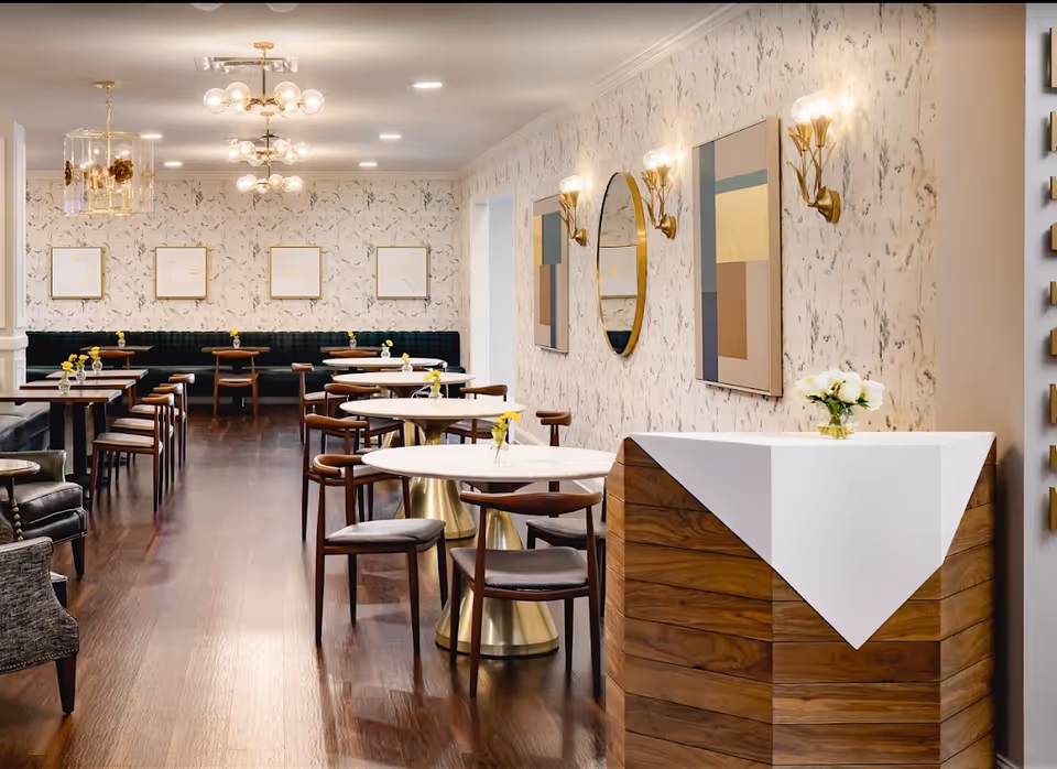 A modern dining room with multiple round and rectangular tables, each with small vases holding yellow flowers. The room features dark wooden chairs, a dark green cushioned bench along the back wall, and light floral wallpaper. There are three gold and glass ceiling light fixtures, two gold wall sconces with lights, and abstract artwork and a round mirror on the walls. A wooden reception desk with a white cloth and a small vase of white flowers is visible in the foreground.