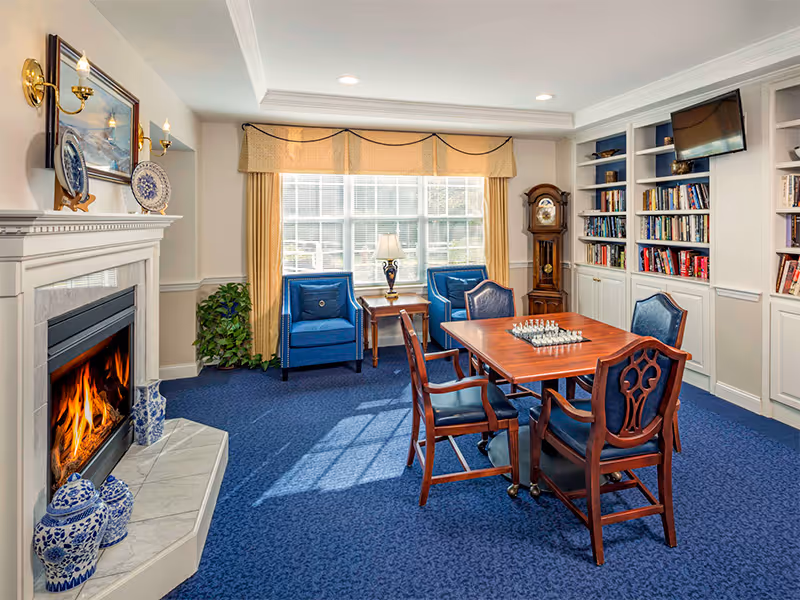 Sunlit common room featuring a lit fireplace, blue upholstered chairs, a wooden game table with a chess set, and built-in bookshelves.