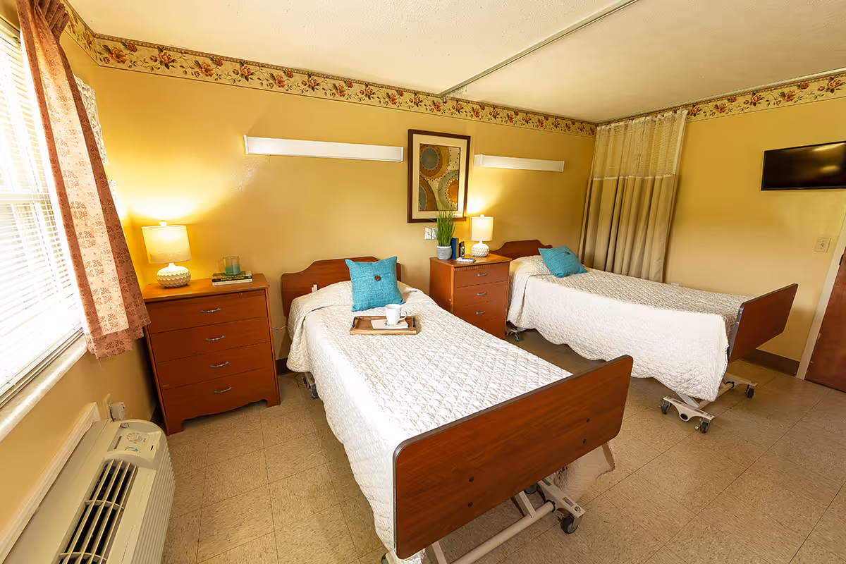 A bright room with two hospital-style beds, each with white quilted bedspreads and turquoise pillows. Between the beds are two wooden nightstands with lamps, books, and a plant. A framed abstract artwork hangs on the wall above the nightstands. The room has a window with floral curtains, a wall-mounted TV, and a beige curtain divider. The floor is tiled, and there is an air conditioning unit below the window.