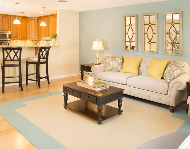 Bright living room with a beige sofa, decorative pillows, coffee table, and an open kitchen with bar stools.