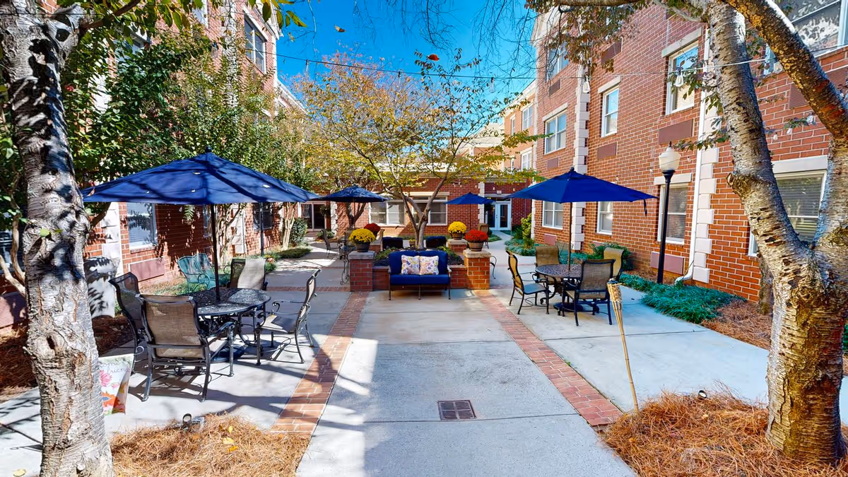 Outdoor courtyard area at Dunwoody Place with brick buildings surrounding a paved patio. The patio features several tables with blue umbrellas and chairs, a central seating area with a blue cushioned bench, and trees providing shade. The sky is clear and blue.