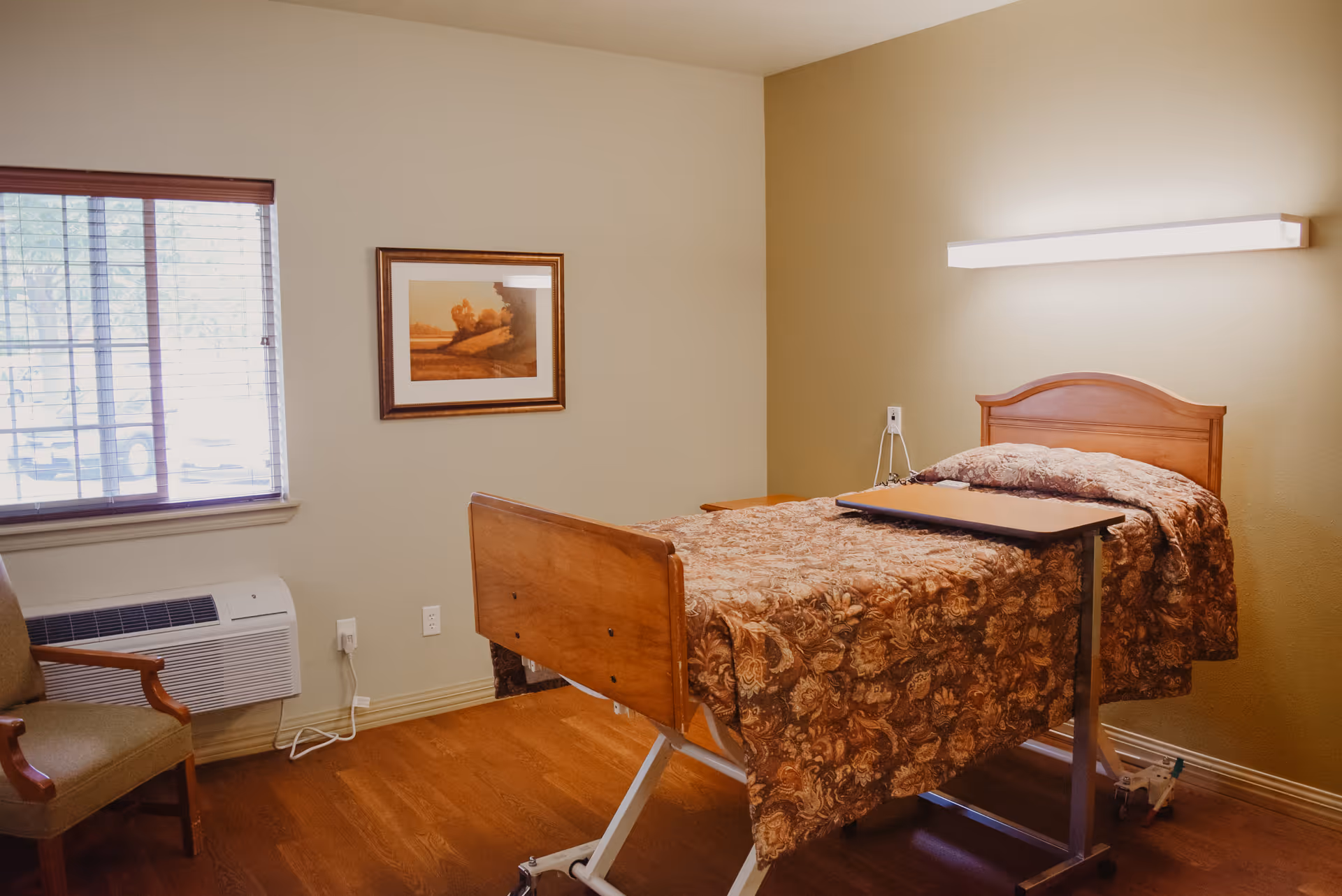 A simple, clean bedroom in a care facility with a single bed covered in a brown floral bedspread, a wooden chair, a window with blinds, a wall-mounted light above the bed, and a framed landscape painting on the wall.
