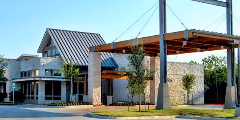 Exterior view of a modern building with a stone facade and a metal roof, featuring a covered entrance supported by stone pillars and surrounded by small trees and landscaping.