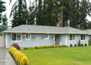 Single-story blue-gray facility front with windows, hanging planters, and a manicured lawn.