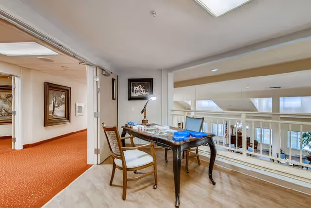 A small seating area with a wooden table and two chairs is situated near a railing overlooking a lower floor. The table has various papers and a desk lamp on it. The floor is light wood, and the adjacent hallway has orange carpet and framed artwork on the walls.