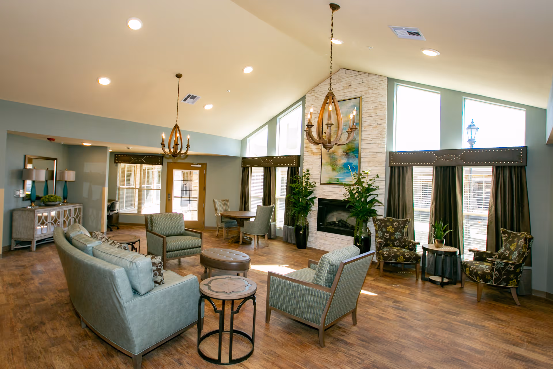 Bright communal living room with sofas and armchairs arranged around a fireplace, chandeliers, and large windows.