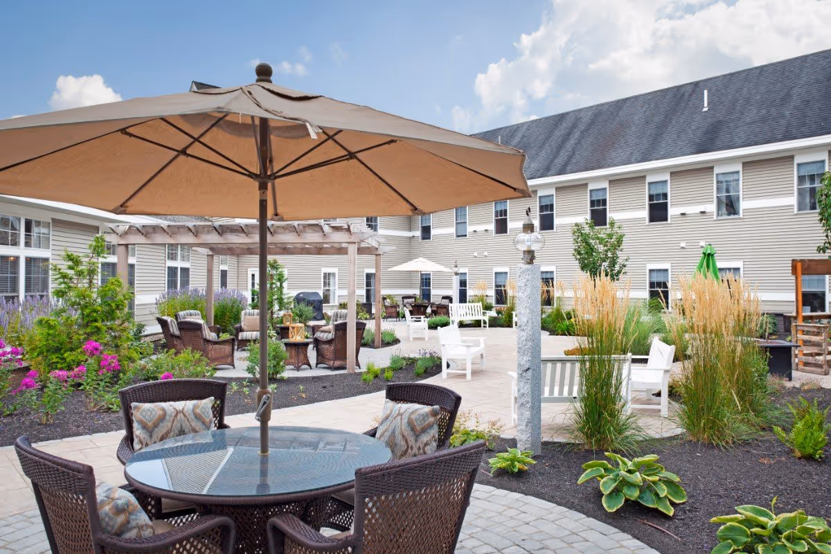 Outdoor courtyard patio at Huntington Common with an umbrella-shaded table, wicker chairs, landscaped plantings and the facility building in the background.