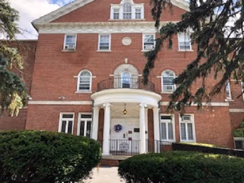 Brick building with arched entrance, white columns, and hedges at the front walkway.