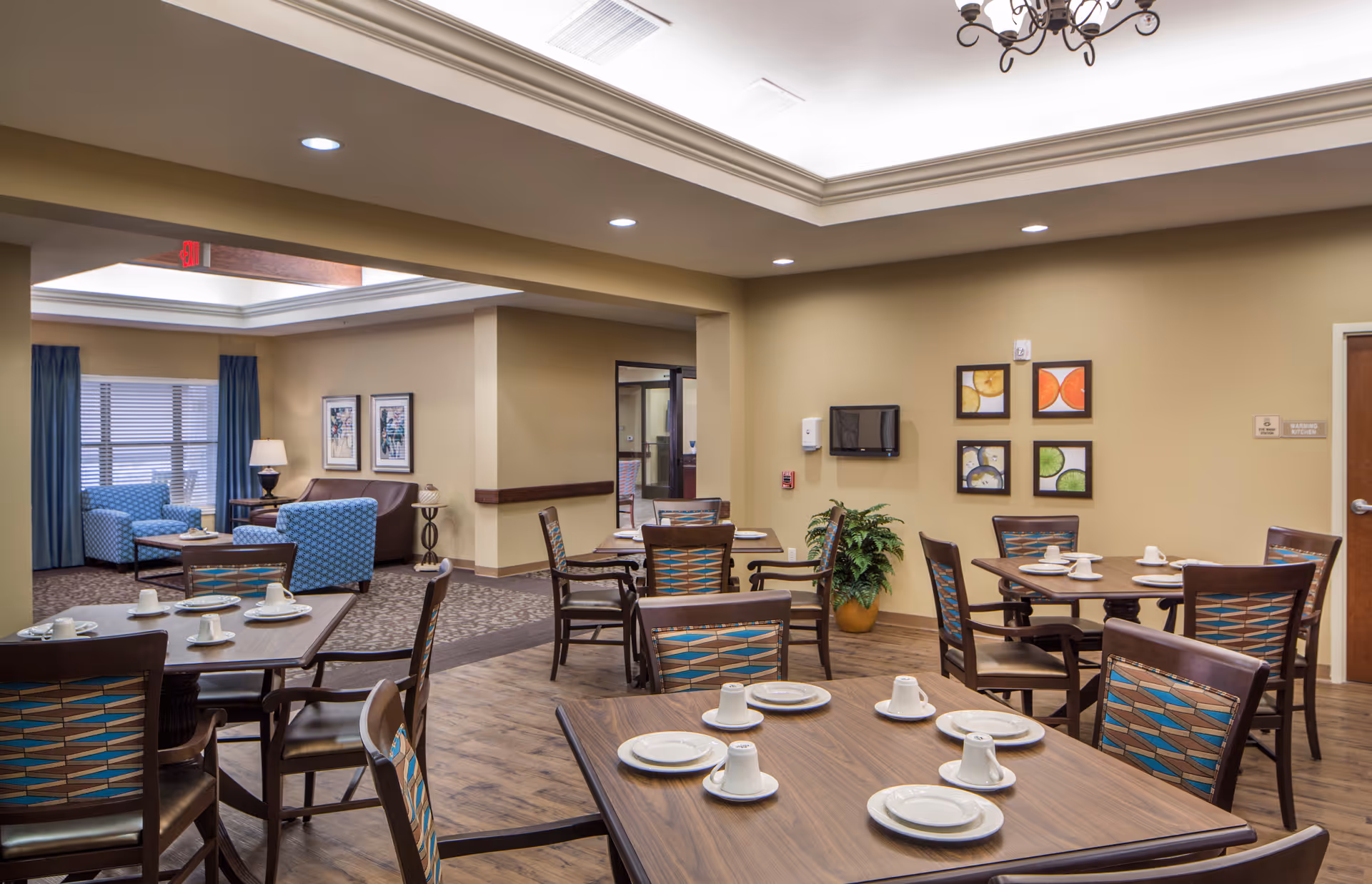 A dining area in a senior living facility with several wooden tables set with white plates and cups. The chairs have patterned upholstery. In the background, there is a seating area with blue armchairs and a brown sofa, along with framed artwork on the walls and a potted plant. The room has beige walls, recessed lighting, and a chandelier.