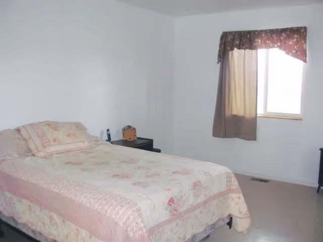A simple bedroom with a bed covered in a floral and striped quilt, a pillow, a small nightstand with a phone and a decorative item, and a window with a brown curtain partially drawn.