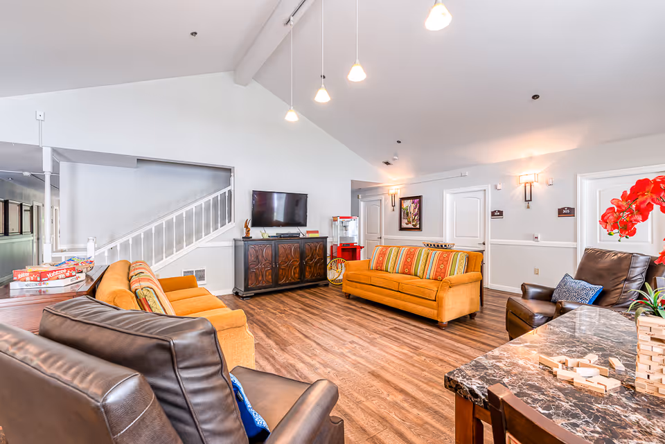 A bright and spacious living room area in a senior living facility featuring two orange sofas with striped cushions, two dark brown leather armchairs, a flat-screen TV mounted on a wooden cabinet, a popcorn machine, and a table with a Jenga game and red flowers. The room has wood flooring, white walls, and pendant lights hanging from a vaulted ceiling.