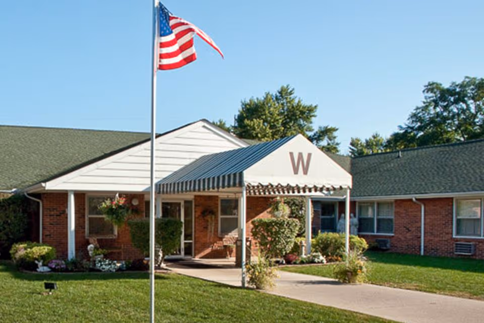 Exterior view of The Woodlands facility showing a single-story brick building with a green roof and a covered entrance marked with a large letter 'W'. An American flag is flying on a flagpole in front of the building, surrounded by well-maintained grass, shrubs, and trees under a clear blue sky.