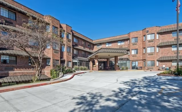 Brick senior living building with a covered entrance and wide driveway, clear blue sky.