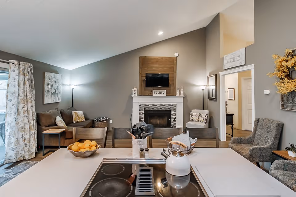 A cozy living room with a vaulted ceiling painted in gray tones. The room features a white fireplace with a mounted TV above it and a wooden accent panel. There are two floor lamps on either side of the fireplace, a brown sofa with cushions on the left, and two patterned armchairs on the right. A kitchen island with a stovetop, a white kettle, cooking utensils, and a bowl of oranges is in the foreground. The room has a warm and inviting atmosphere with decorative elements like a floral painting, curtains, and a wreath on the wall.