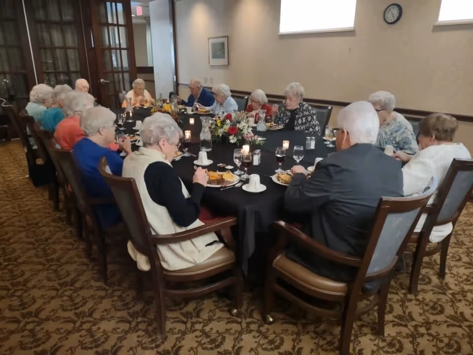 A group of elderly people sitting around a large rectangular dining table covered with a black tablecloth, eating a meal together in a well-lit room with carpeted floor and beige walls. The table is set with plates of food, glasses of water and wine, candles, and a floral centerpiece.