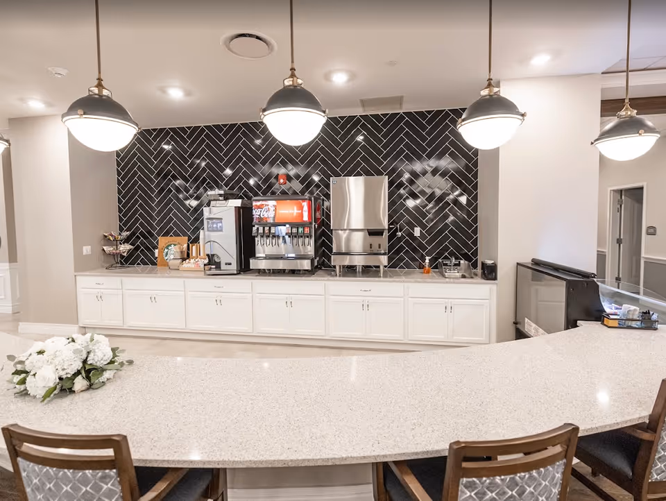 A modern beverage station with a soda fountain, coffee machine, and ice dispenser set against a black herringbone tile backsplash. The countertop is light-colored with a bouquet of white flowers placed on it, and there are three pendant lights hanging from the ceiling. Several chairs with patterned backs are positioned in front of the counter.