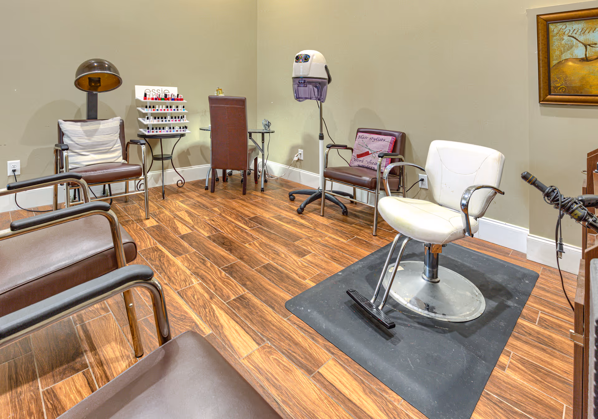 Interior view of a salon area in an assisted living facility featuring a white salon chair on a black mat, several brown chairs, a hair dryer hood, a nail polish display rack, and a small table with a brown chair. The room has wood-patterned flooring and beige walls with a framed picture.