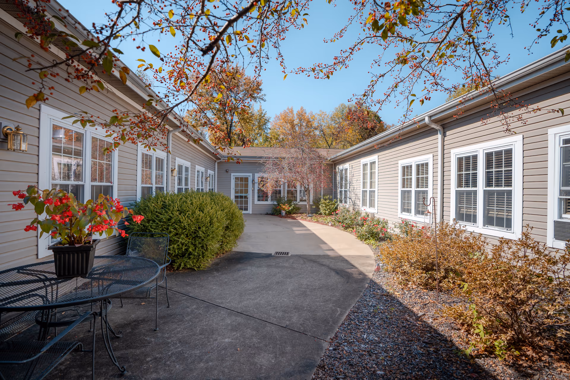 Outdoor courtyard area of a senior living facility with beige siding buildings on both sides, multiple white-framed windows, a concrete walkway, bushes, and a metal table with chairs and a potted plant with red flowers. Trees with autumn foliage are visible in the background under a clear blue sky.
