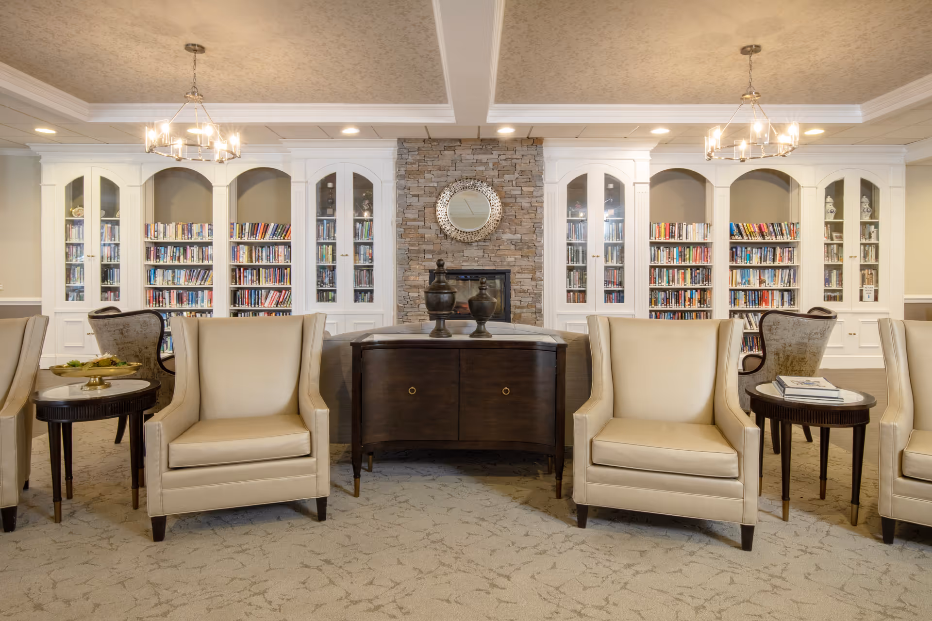A cozy and elegant common area featuring beige armchairs and small round tables arranged in front of built-in white bookshelves filled with books. A stone fireplace with a round decorative mirror above it is centered between the bookshelves. The ceiling has recessed lighting and two modern chandeliers.