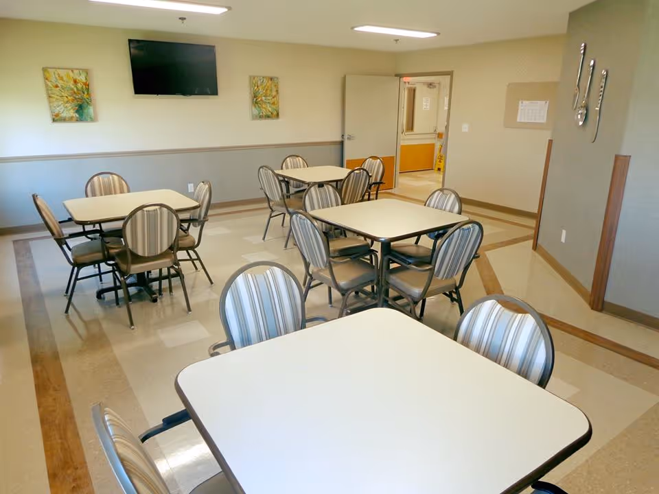 A clean and well-lit dining room with several square tables and striped cushioned chairs arranged neatly. The room has beige walls with two colorful paintings and a flat-screen TV mounted on one wall. There is an open door leading to a hallway, and the floor has a patterned design with a mix of beige and brown tones.