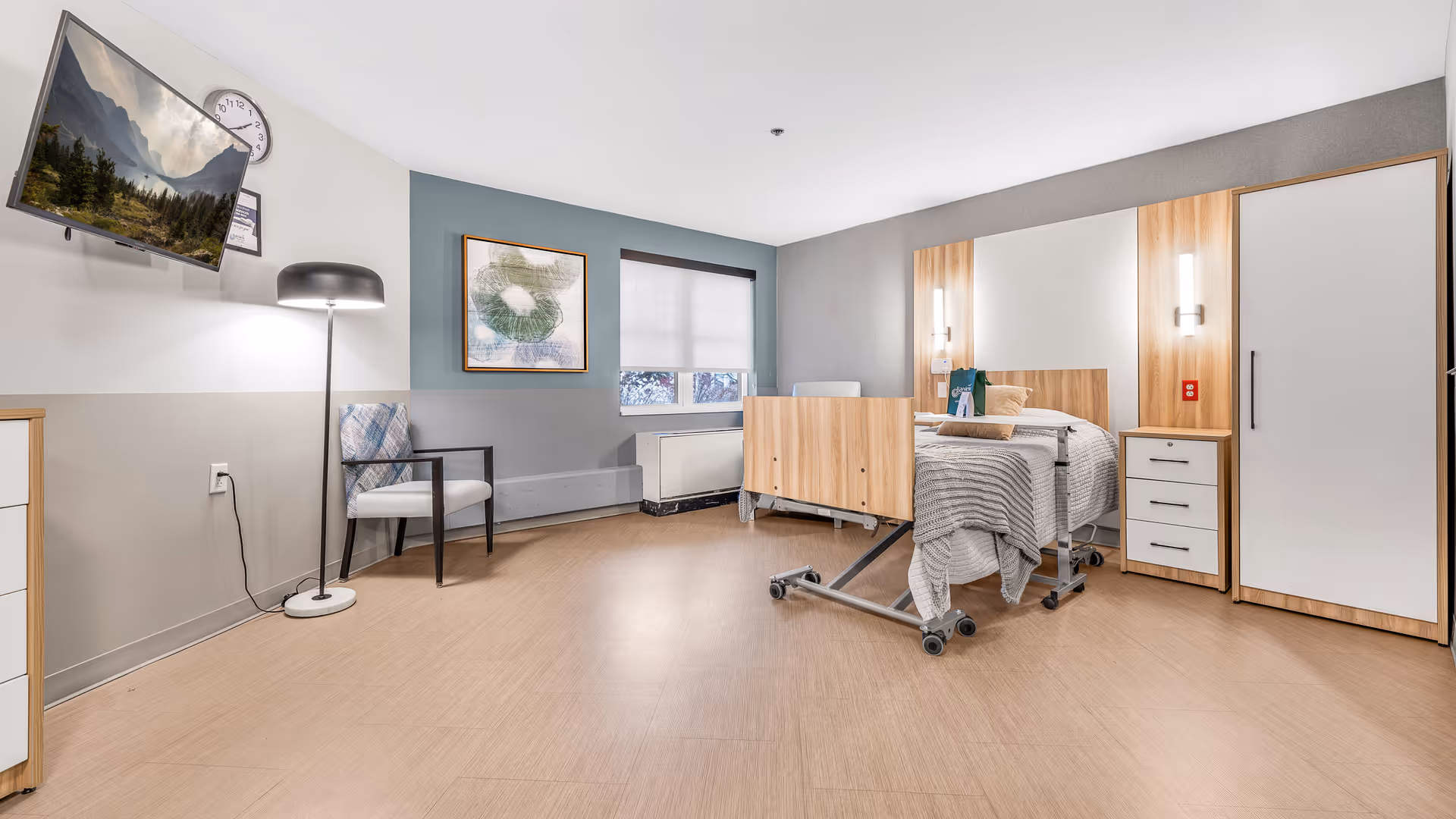 A bright and spacious rehabilitation room featuring a hospital bed with wooden accents, a bedside table with drawers, a wardrobe, a chair with a patterned cushion, a floor lamp, a wall-mounted TV displaying a nature scene, a clock, and a large window with a white roller shade.