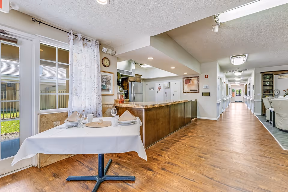 Interior view of a senior living facility showing a small dining table set for two with white tablecloth, napkins, glasses, and plates near a glass door with sheer curtains. To the right is a long hallway with wooden flooring and white walls, leading to other rooms. A kitchen counter with a granite top and stainless steel refrigerator is visible in the background.