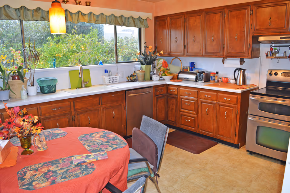 Sunlit kitchen with wooden cabinets, stainless steel stove and dishwasher, a table with a red tablecloth, and a large window over the sink.