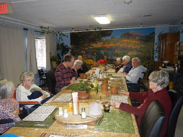 A group of elderly residents seated around a long communal dining table in a decorated dining room.