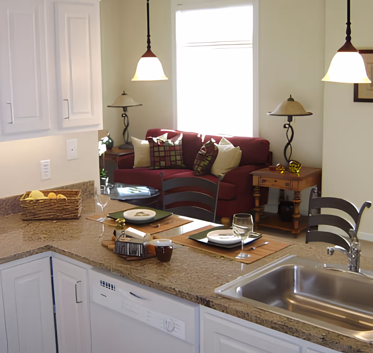 View of a kitchen counter with a sink and two place settings with plates, glasses, and utensils. Behind the counter is a living area with a red sofa adorned with multiple pillows, two side tables with lamps, and a large window letting in natural light.