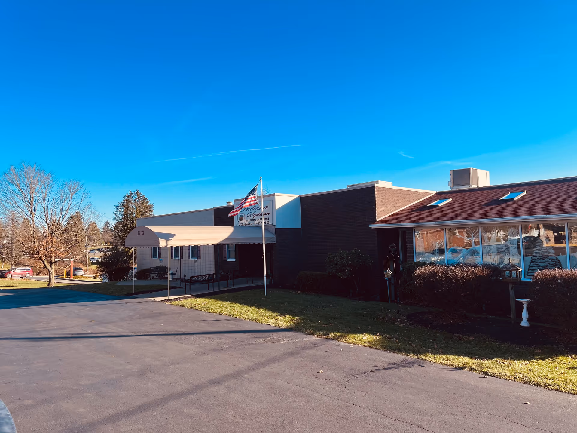 Exterior view of a single-story brick building with a beige awning over the entrance, an American flag on a flagpole, and a clear blue sky. The building has large windows on the right side and some bushes in front. There is a paved driveway and some trees without leaves in the background.