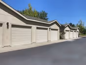 A row of beige garages with closed garage doors along a paved driveway, with trees and blue sky in the background.