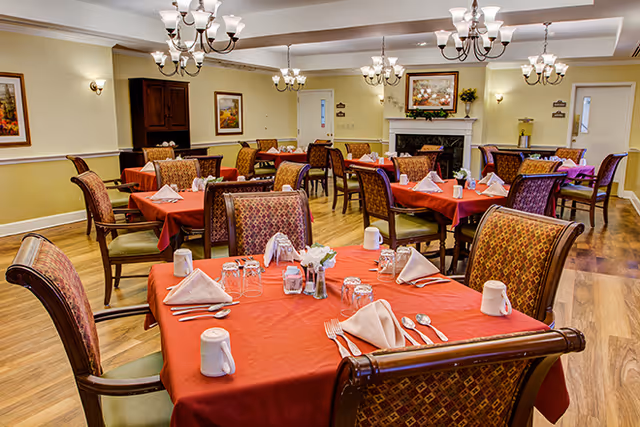 A bright senior living dining room with multiple tables covered in red tablecloths, set with napkins, cups, and utensils.