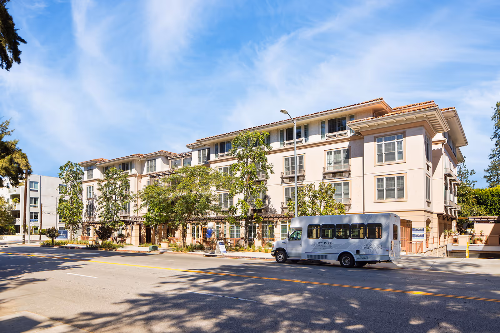 Front exterior of a multi-story senior living building with trees and a white shuttle bus parked on the street.
