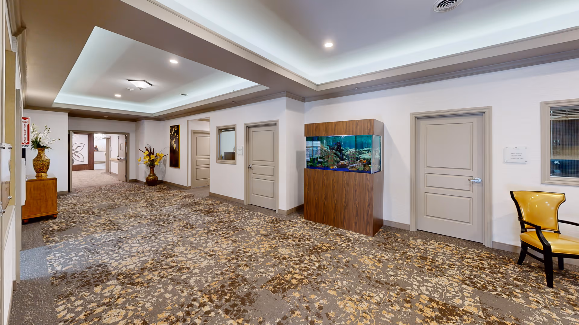 Bright assisted living facility corridor with patterned carpet, a large aquarium against the wall, seating, decorative vases and several doors.