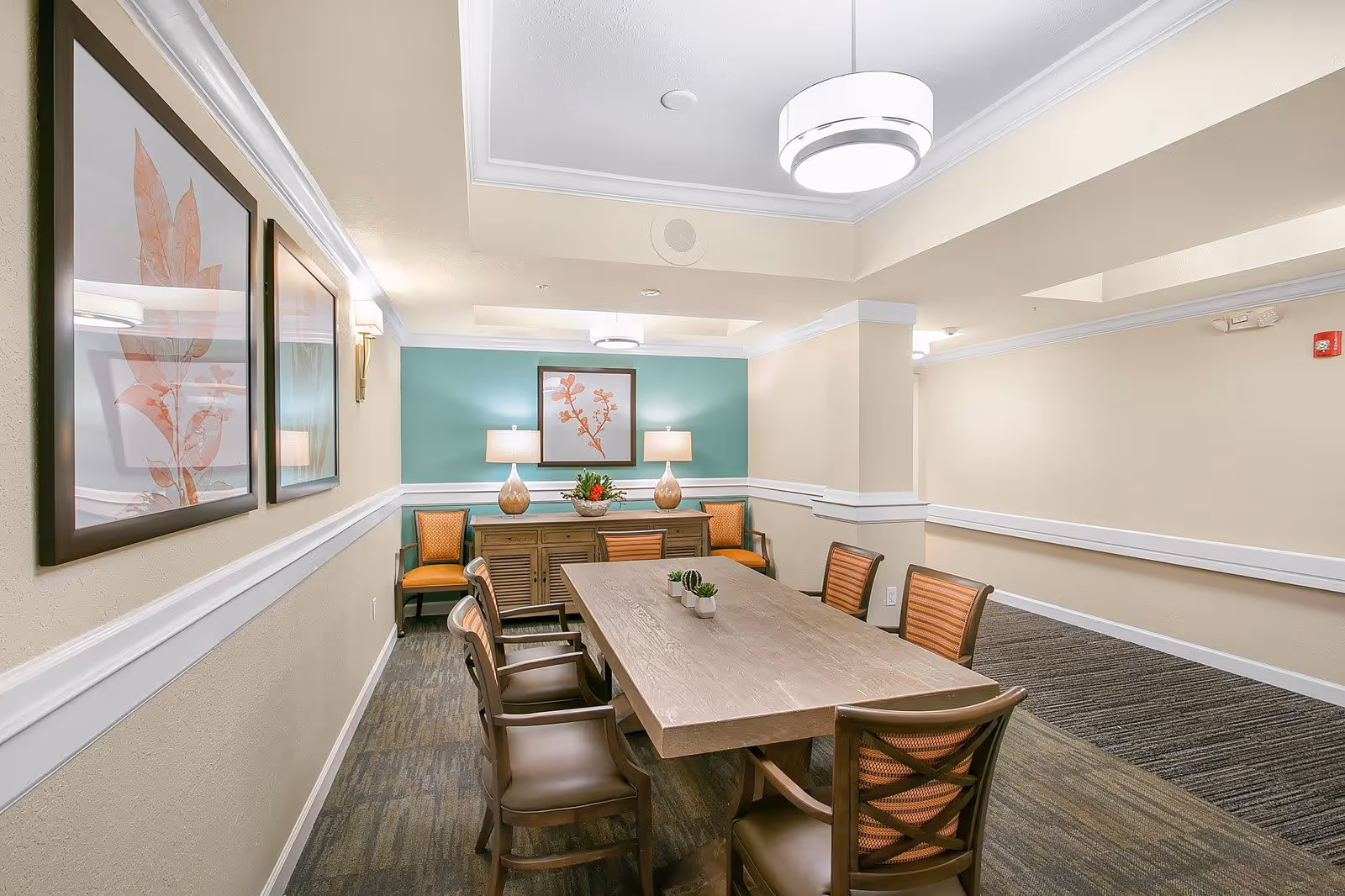 A well-lit dining room with a rectangular wooden table surrounded by eight chairs. The walls are painted beige with a teal accent wall at the far end, decorated with framed botanical artwork and two table lamps on a wooden sideboard. The room has modern ceiling lights and carpeted flooring.