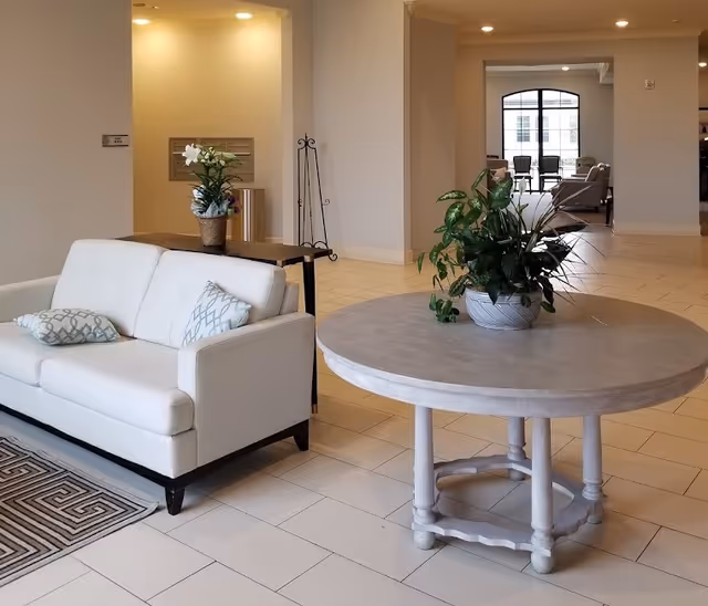 A bright and spacious interior common area with a white sofa adorned with two patterned pillows, a round wooden table with a potted green plant, and a patterned rug on the tiled floor. In the background, there is another seating area with chairs near large windows letting in natural light.