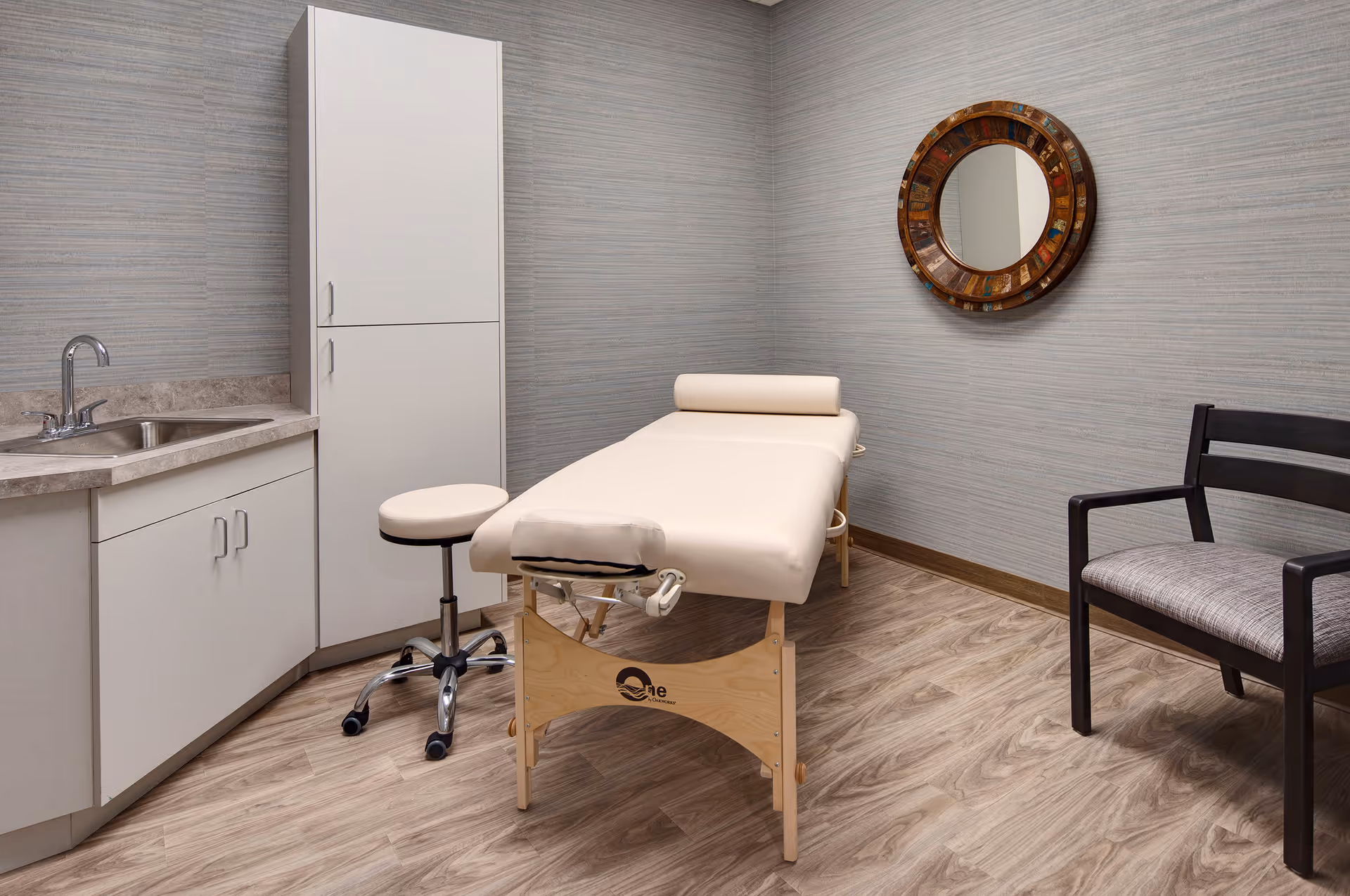 A small treatment room with a padded massage table, a rolling stool, a wooden chair with a cushioned seat, a sink with countertop and cabinets, and a round decorative mirror on the wall.