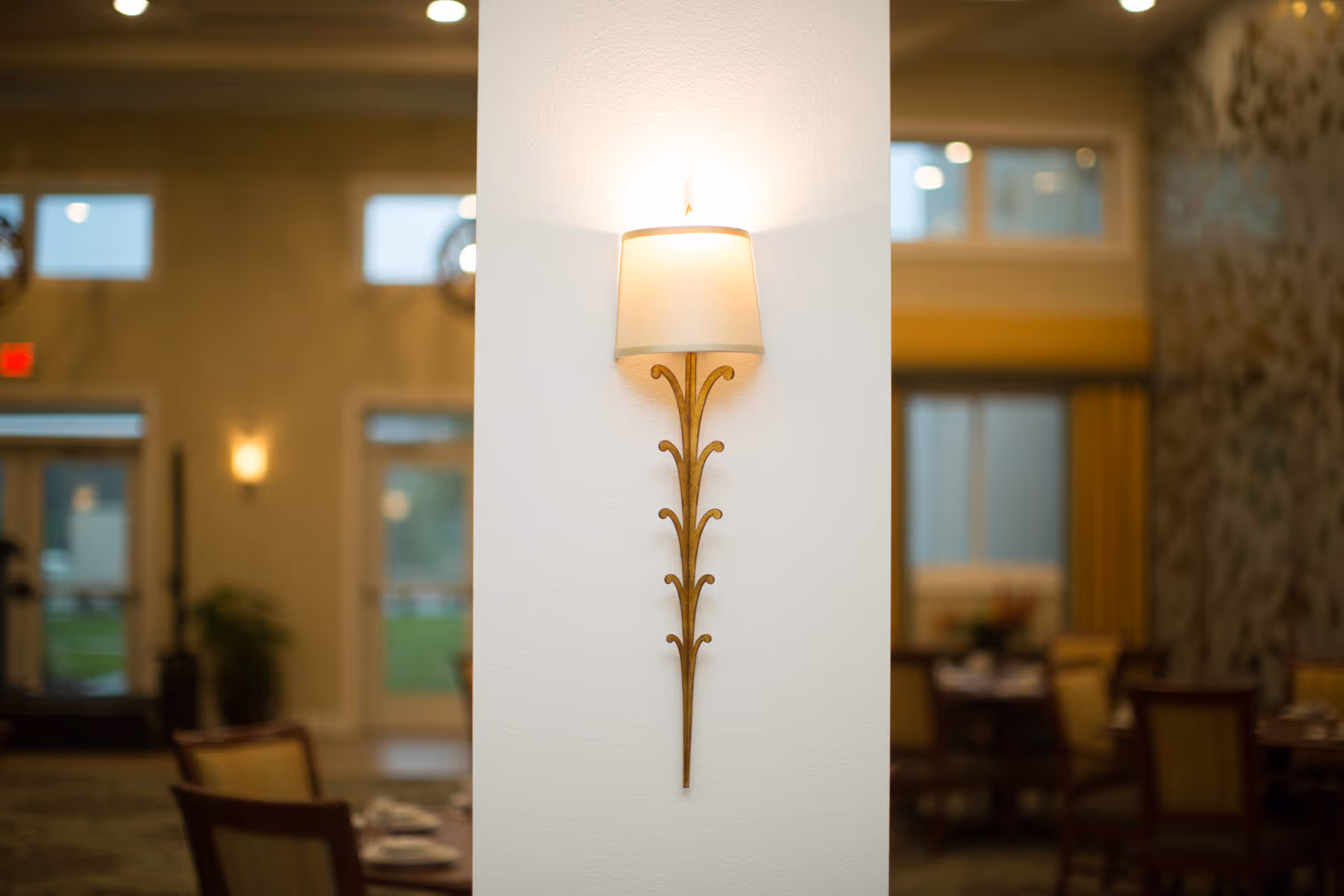 A decorative wall sconce with a white lampshade mounted on a white pillar in a softly lit dining area with tables and chairs in the background.