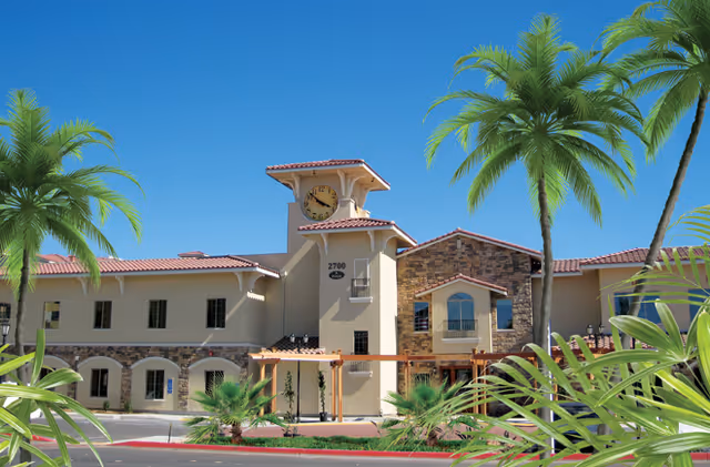 Exterior view of a senior living facility named Cedars @ Paradise Village featuring a two-story building with a clock tower, stone and stucco facade, surrounded by palm trees and landscaping under a clear blue sky.