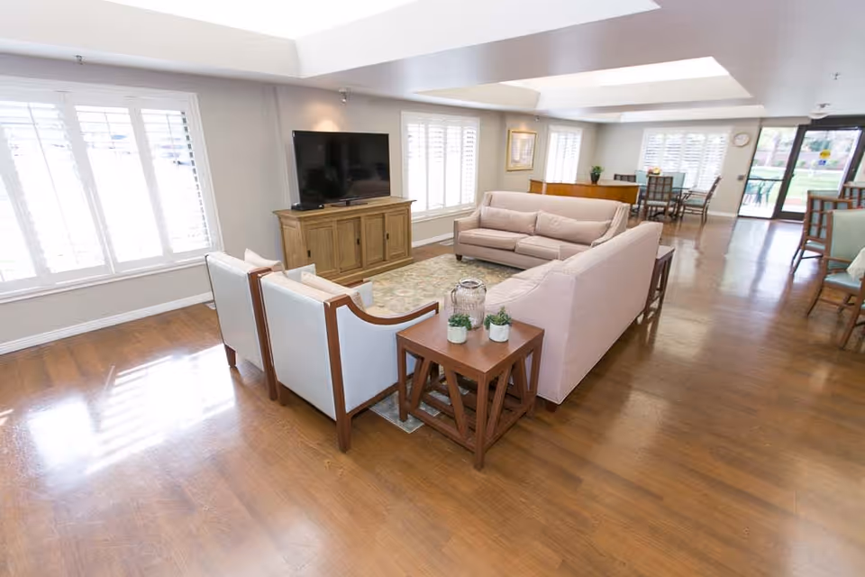 A bright and spacious senior living facility common area with wooden floors, two beige sofas, two white armchairs, a wooden TV stand with a flat-screen TV, and a small wooden side table with potted plants. Large windows with white shutters allow natural light to fill the room. In the background, there is a dining area with tables and chairs and a glass door leading outside.