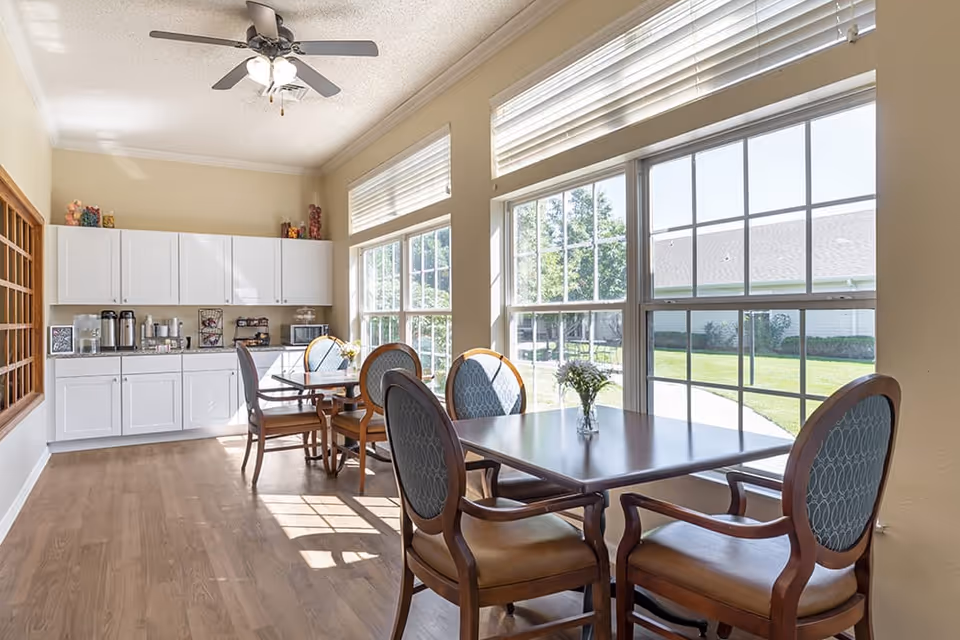 Sunlit dining area with wooden tables and upholstered chairs beside large windows and a small kitchenette with white cabinets.