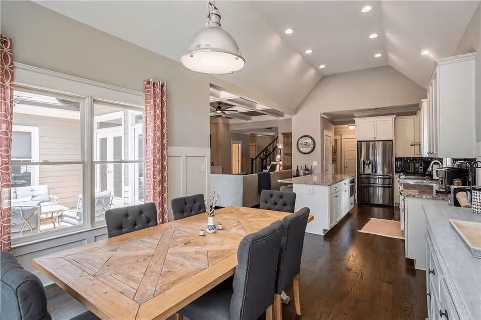 Bright open dining area with a wooden table and upholstered chairs adjacent to a modern kitchen with white cabinets and stainless steel appliances.