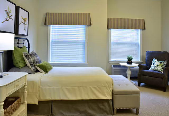 A bright bedroom with a neatly made bed, upholstered bench at the foot, armchair, side table, and two windows with blinds.