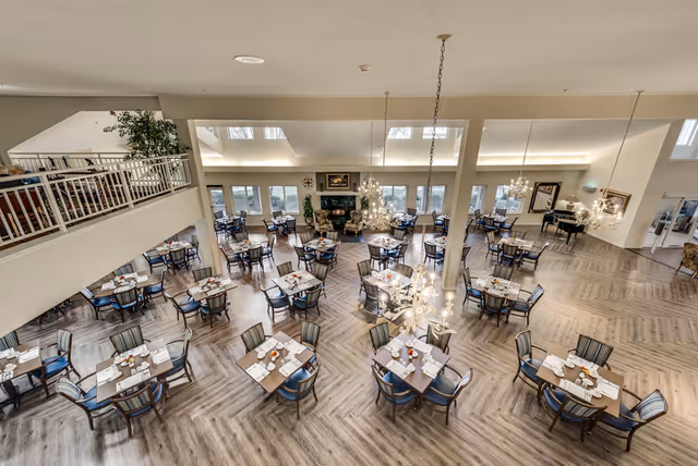 Spacious dining room with multiple tables set with plates, glasses, and napkins. The room features large windows allowing natural light, chandeliers hanging from the ceiling, and a fireplace with seating area in the background. There is a piano on the right side and a mezzanine level with railing and plants on the left.