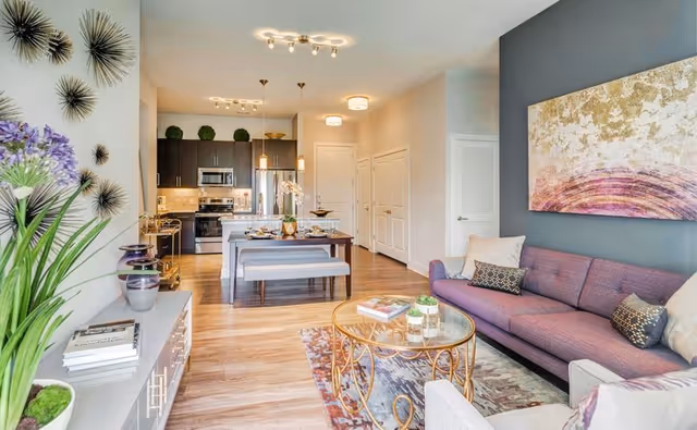 A modern open-concept living and dining area in a senior living facility. The living room features a purple sofa with decorative pillows, a glass coffee table with gold metal legs, and a patterned rug. The dining area has a wooden table with a bench and chairs, set with plates and glasses. The kitchen in the background has dark cabinets, stainless steel appliances, and pendant lighting. Decorative plants and wall art add a cozy touch to the space.