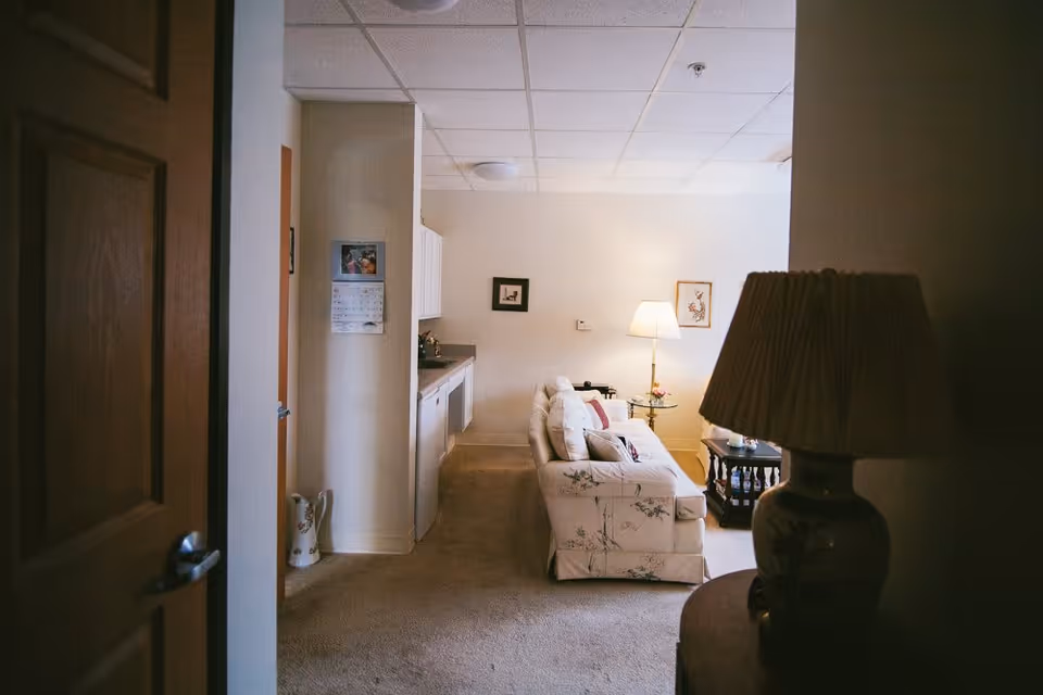 View into a cozy living room area from a hallway, featuring a floral-patterned sofa, a side table with a lamp, framed artwork on the walls, and a small kitchenette with white cabinets on the left side.